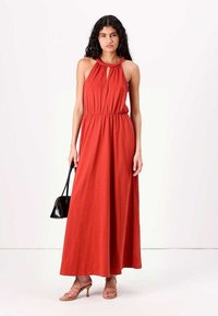 Vestido maxi vermelho com decote halter e cintura elástica, apresentando uma textura suave. Usado com sandálias de tiras e segurando uma pequena bolsa preta.