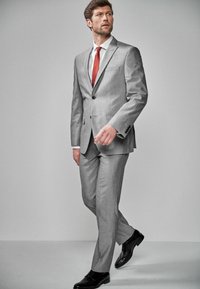 Szürke öltöny, testre szabott fazonban, két gombbal, hajtókás gallérral, és finom textúrával. Fehér inggel és piros nyakkendővel párosítva; fekete elegáns cipőkkel.