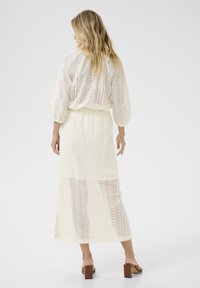Robe longue de couleur crème avec des motifs en dentelle complexes, manches mi-longues et taille cintrée, présentant un design fluide et une fente sur le côté.