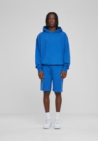 Blauwe katoenen hoodie met een zak aan de voorkant, gecombineerd met bijpassende blauwe shorts. Het model draagt witte sneakers en witte sokken tot de enkel.