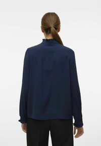Marineblauwe blouse met een gestructureerde afwerking, gerimpelde kraag en lange mouwen. Voorzien van knoopmanchetten en een relaxte pasvorm, gedragen met zwarte broeken.
