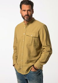 Camicia a maniche lunghe verde oliva in misto lino con colletto con bottoni, tasca sul petto e bottoni frontali. Indossata con jeans blu sbiaditi.