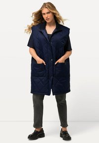 Donna con lunghi capelli biondi, che indossa un gilet senza maniche blu scuro trapuntato, una maglietta nera, jeans grigi e mocassini neri, in piedi con le mani nelle tasche.