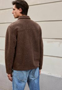 Veste en mélange de laine brun avec un col, présentant une finition texturée. Le modèle l'associe à un jean en denim bleu clair.