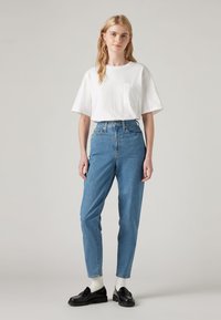 Ung kvinna med långt blont hår som bär en vit t-shirt, högmidjade blå jeans, vita strumpor och svarta loafers, stående mot en enfärgad bakgrund.