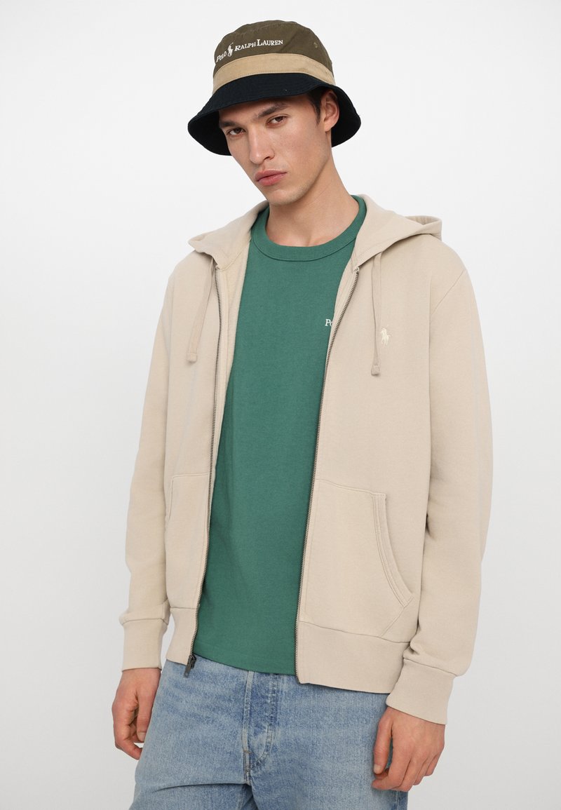 Beige zip-up hoodie met een kangoeroezak, gecombineerd met een groene T-shirt met ronde hals en lichtblauwe jeans. Het model draagt ook een bucket hat.