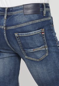 Tmavomodré džínsy z denimu s dizajnom piatich vreciek, viditeľným prešívaním a kontrastnou koženou nášivkou na páse. Opotrebovaný vzhľad na vreckách.