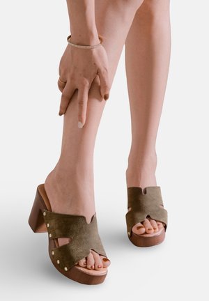 Femme portant des talons carrés marron à bout ouvert avec des détails cloutés, une main reposant sur sa jambe inférieure contre un fond blanc uni.
