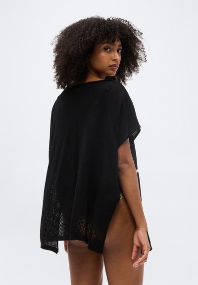 Γυναίκα με κατσαρά μαλλιά που φοράει μαύρο πλεκτό ποντσο, γυρισμένη προς τα πίσω, κοιτάζει από πάνω στον ώμο της μπροστά σε απλό φωτεινό φόντο.