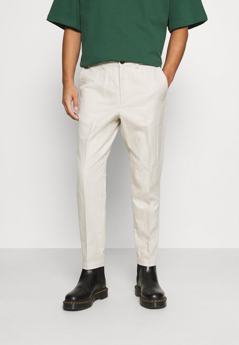 Pantalon beige ajusté, confectionné dans un tissu léger. Il possède des poches latérales, une fermeture par bouton, et une texture lisse. Bottines noires.