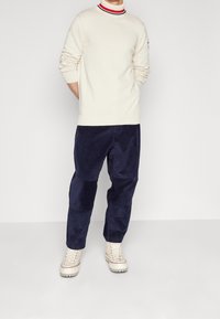 Gestrickter cremefarbener Pullover mit geripptem Kragen, kombiniert mit marineblauen Cordhosen. Die Schuhe sind hellbeige mit einer strukturierten Sohle und roten Akzenten.