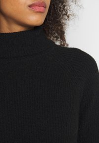 Pull en tricot noir avec un col roulé côtelé haut. La texture est douce et épaisse, avec une coupe ajustée qui met en valeur le décolleté.