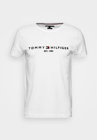 Bílé krátké tričko s krátkým rukávem s textem "Tommy Hilfiger EST. 1985" a značkovým logem v červené, bílé a modré barvě umístěným na hrudi.