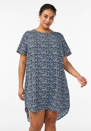 Kvinde iført en kortærmet mørkeblå kjole med hvidt blomstermønster, stående mod en ensfarvet hvid baggrund.