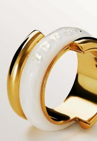 Brazalete dorado y blanco con una superficie lisa y brillante, con acentos claros y un diseño curvado, que muestra una mezcla de materiales y texturas.