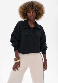 Czarna, krótka kurtka z dzianiny z kołnierzykiem, z dwiema kieszeniami na piersi i zapięciem na guziki. W zestawieniu z jasnymi, dopasowanymi spodniami.