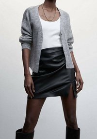 Graue Strickjacke über einem weißen Tanktop, kombiniert mit einem schwarzen Mini-Lederrock mit Seitenschlitz und kniehohen schwarzen Stiefeln.