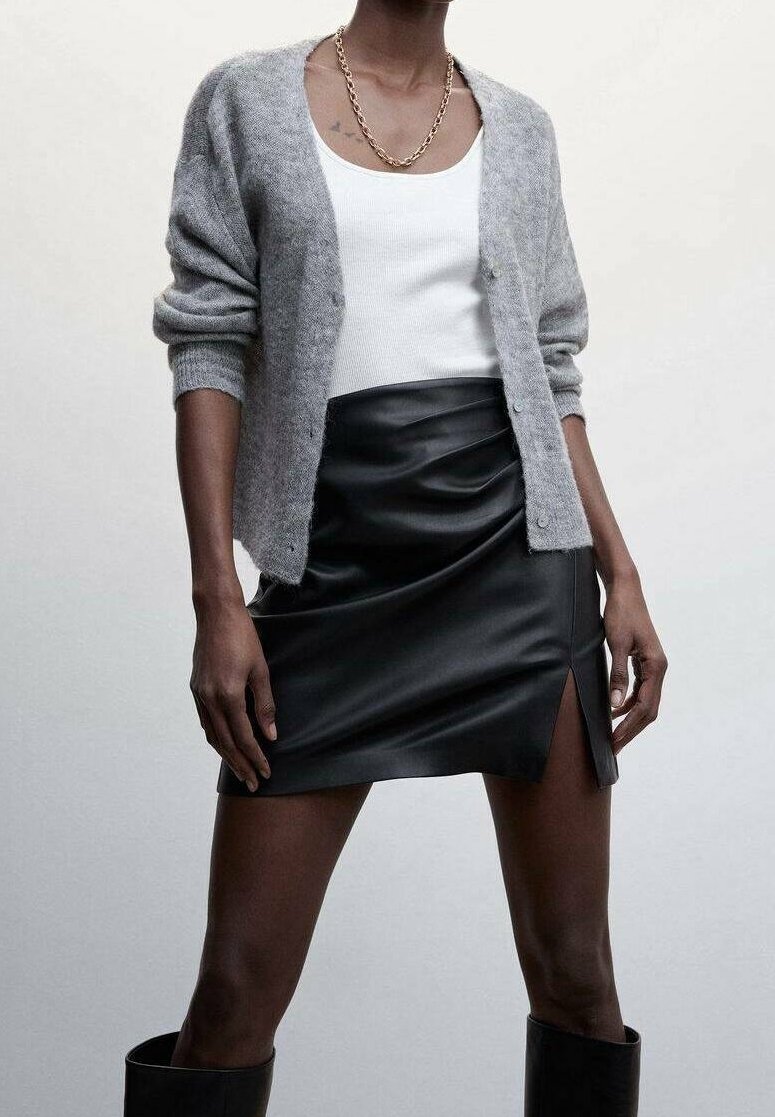 Graue Strickjacke über einem weißen Tanktop, kombiniert mit einem schwarzen Mini-Lederrock mit Seitenschlitz und kniehohen schwarzen Stiefeln.