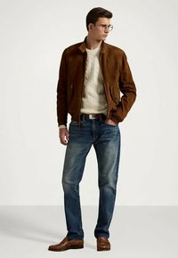 Ung mann iført brun semsket jakke, kremfarget kabelstrikket genser, blå jeans, brune lær-sko og briller, stående med den ene hånden i lomma.