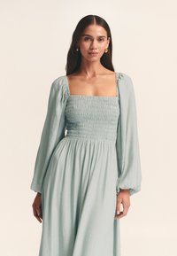 Svetlomodré šaty s naškrobeným vrškom, štvorcovým výstrihom a dlhými nafúknutými rukávmi. Mäkká, textúrovaná látka splýva do voľného strihu v páse.