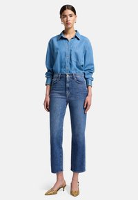 Chemise en denim bleu à boutons avec poche poitrine, associée à un jean droit bleu taille haute et des chaussures à talons jaunes à motifs.