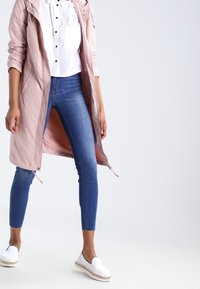 Ljusrosa trenchcoat med dragkedja och huva; ihop med en vit skjorta med knappar, blå skinny jeans och vita slipskor.