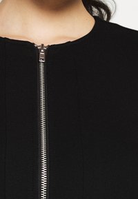 Detail černého oděvu se stříbrným zipem, který je částečně rozepnutý u výstřihu na horní části hrudníku a krku osoby.