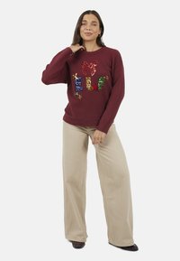 Pull en tricot bordeaux avec un motif "ELF" en sequins et un détail nœud. Associé à un pantalon beige à jambes larges. Coupe décontractée, tissu texturé.