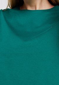 Mujer con una camiseta de cuello redondo en color verde azulado, con clavículas visibles y cabello largo castaño, mostrada desde el cuello hasta la parte superior del pecho.