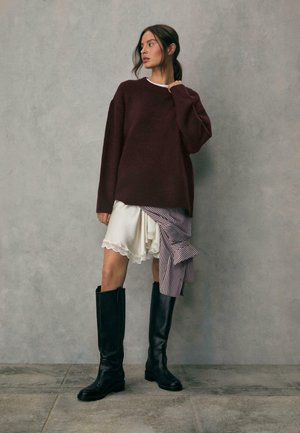 Burgundski oversized pulover, suknja od krem čipkastog obruba, prugasti detalji, i visoke crne kožne čizme. Glatka tekstura, opušten kroj.