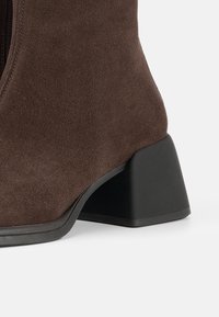 Brown suede ankle boot featuring a textured finish, a zip closure, and a black block heel design.