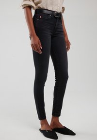 Inimene, kes kannab musti kitsaid teksapükse, sisse söödetud triibulist särki, musta vööd ja musta teravate ninadega madalaid kingi, seisab lihtsal heledal taustal.