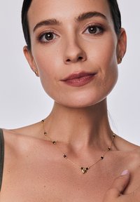 Złoty naszyjnik z czarnymi koralikami, z delikatnym łańcuszkiem i małym zawieszką na środku, noszony na odsłoniętej skórze. Towarzyszą mu proste kolczyki wkrętki.