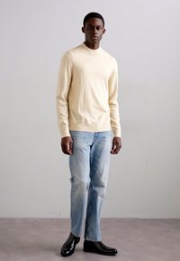Beigefarbener Langarmpullover, lockere Passform, kombiniert mit hellblauen Jeans und glänzenden schwarzen Stiefeln. Einfaches Design, glatte Textur.