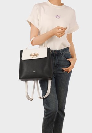 Donna con top bianco a maniche corte e jeans scuri tiene uno zaino in pelle bianco e nero per la cinghia vicino alla vita su uno sfondo chiaro.