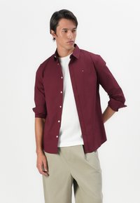 Bordeaux knoopshirt met lange mouwen, witte knoopsluiting en klein logodetail. Draag het over een wit T-shirt en combineer het met beige broek.