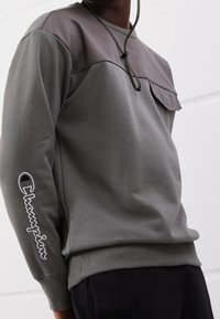 Uomo che indossa un felpa grigia Champion con taschino sul petto e logo sul braccio sinistro, abbinata a pantaloni neri.