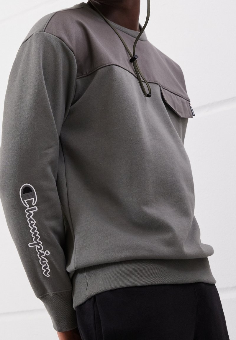 Uomo che indossa un felpa grigia Champion con taschino sul petto e logo sul braccio sinistro, abbinata a pantaloni neri.