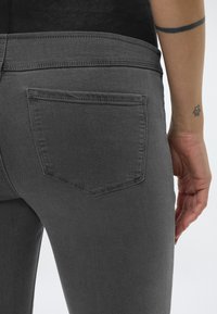 Κοντινό πλάνο ενός ατόμου που φορά σκούρο γκρι τζιν με ορατή πίσω τσέπη, εμφανίζοντας το χέρι και τον πήχη με δύο μικρά τατουάζ.