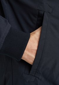 Chaqueta navy con una textura suave, que presenta un bolsillo lateral y puños de canalé. El bolsillo tiene un detalle de solapa triangular en el borde.