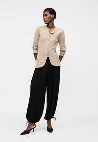 Vrouw in een beige vest met knopen en een gestructureerd patroon, zwarte wijde broek met trekkoord en zwarte puntschoenen, staand met handen achter de rug.