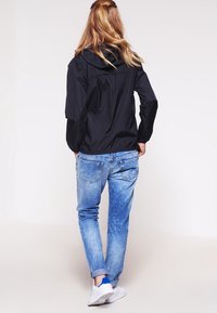 Svart lättviktsjacka med huva, tillsammans med ljusblå jeans. Jackan har elastiska manschetter och en mjuk yta. Vita sneakers synliga.