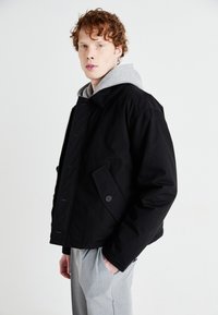 Black bomber jacket with a structured design, featuring a buttoned front, side pocket, and a grey hoodie underneath. Textured fabric, relaxed fit.