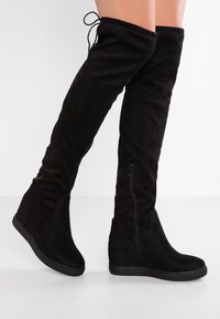 Bottes noires montantes au-dessus du genou en daim souple avec semelle compensée. Elles possèdent une fermeture éclair latérale et des lacets ajustables en haut.