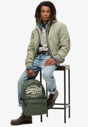 Groene vintage rugzak met wit logo, gedragen door een persoon in een olijfgroene jas, grijze sweater en blauwe spijkerbroek, zittend op een zwart krukje. Bruine laarzen.