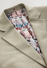 Blazer beige texturizado con cuello de muesca, que revela un forro con estampado floral en suaves colores pastel de rosa, azul y marrón.