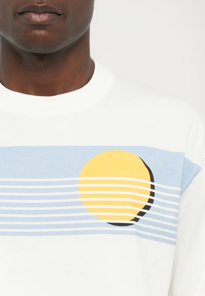 Tricou alb din bumbac cu o bandă orizontală albastră, decorată cu un cerc galben și un semicerc negru, formând un design grafic al soarelui.