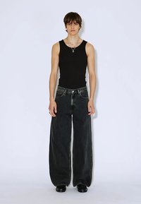 Musta toppi yhdistettynä leveälahkeisiin mustiin farkkuihin. Kankaan rakenne on teksturoitu ja siinä on kontrastikirjailut. Malli seisoo yksinkertaista taustaa vasten.