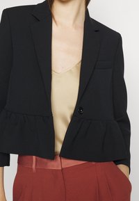 Le modèle porte un blazer noir court avec un ourlet à volants sur un top beige, assorti à un pantalon rouge, illustrant une tenue moderne et élégante.