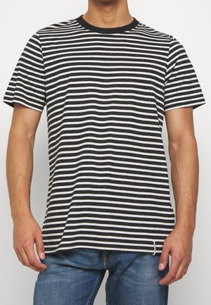 Mann trägt ein kurzärmeliges T-Shirt mit schwarzen und weißen horizontalen Streifen und dunklem Kragen sowie blaue Jeans, steht vor einem schlichten weißen Hintergrund.